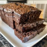 A stack of three delicious chocolate brownies sits on a dark gray surface, surrounded by chocolate chips.