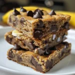 Four chocolate chip banana bread bars on a wooden cutting board, next to a bowl of chocolate chips and a banana.